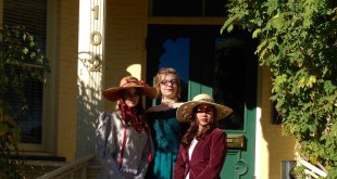 Three young women in period dresses and hats stand on the front steps of the Rinckel Mansion.